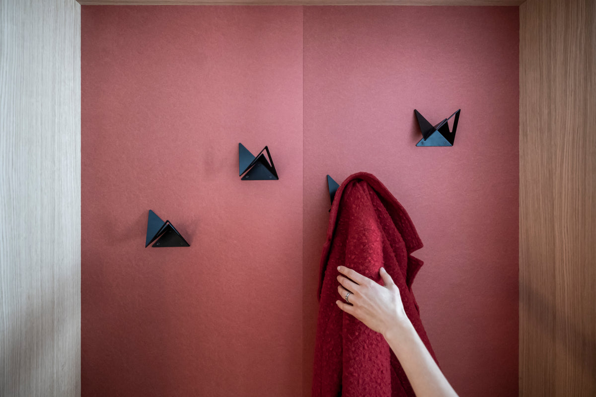 Schwarze Wandhaken Modell Flock in geometrischer Dreiecksform auf einer altrosafarbenen Wand im Hotel Garni Reider, ein roter Mantel wird gerade aufgehängt.