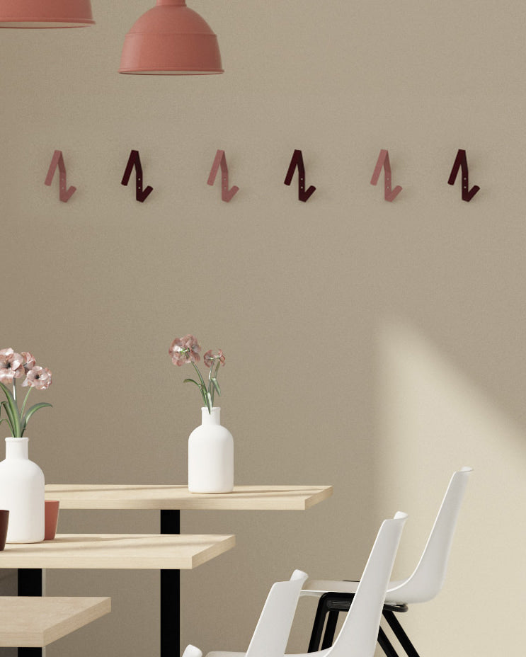 Reihe von modernen, z-förmigen Garderobenhaken in verschiedenen Rosé- und Rottönen an einer beigen Wand in einem hellen Restaurant mit Holztischen und weißen Stühlen.