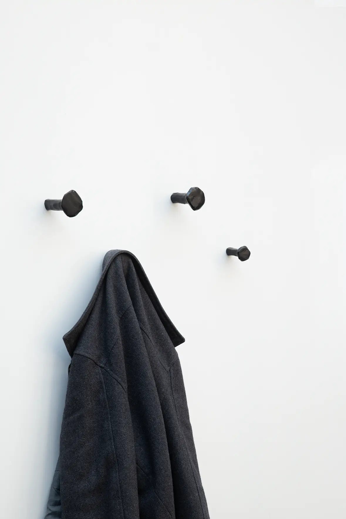 Vier schwarze Schmiedenägel an einer weißen Wand, wobei an einem der Haken eine dunkle Jacke aufgehängt ist.