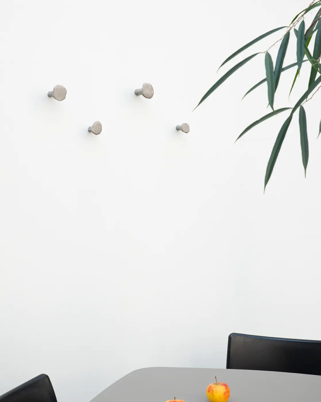 Ein Set aus vier grauen Schmiedenageln in unterschiedlichen Größen, unregelmäßig an einer weißen Wand über einem Tisch mit Äpfeln und Zweigen angeordnet.