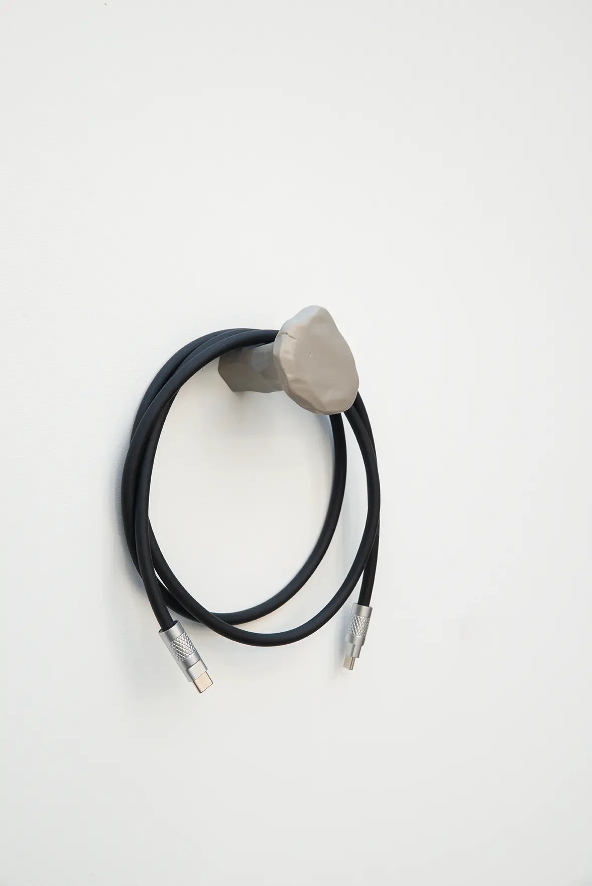 Ein handgefertigter, grauer Schmiedenagel bzw. Stifthaken von Florian Knöbl montiert an einer weißen Wand, um den ein schwarzes Kabel mit Metallsteckern geschlungen ist.