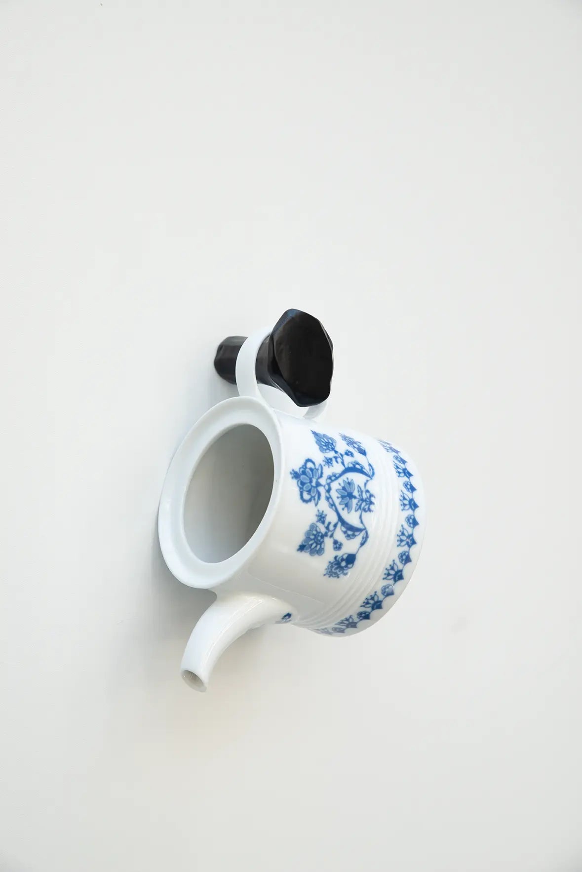 Ein schwarzer Schmiedenagel als Wandhaken, an dem eine weiße Teekanne mit blauem Zwiebelmuster-Dekor am Griff aufgehängt ist.